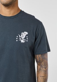 Čovjek obučen u tamnoplavu majicu s bijelim likom sokola i šest zvjezdica na prsima, pokazuje tetovirani desni podlakticu s ružama i vage.