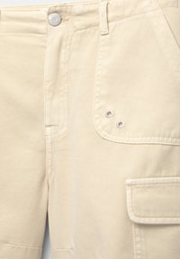 Beige bomullscargobyxor med stora sidfickor, stickade sömmar, metallknappstängning och dekorerande metallöglor. Slät textur.