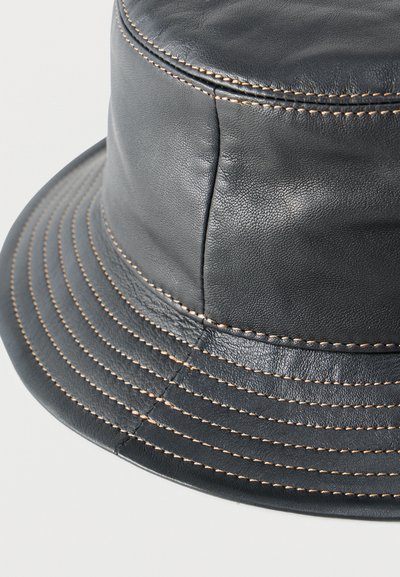 Καπέλο τύπου bucket από μαύρο δέρμα με μπεζ ραφές γύρω από το πάνω μέρος και το γείσο σε απλό ανοιχτό φόντο.