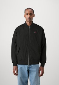 Svart bomberjakke laget av nylon, med glidelås foran, ribbede mansjetter, sidelommer og en liten logo på venstre bryst. Glatt tekstur.
