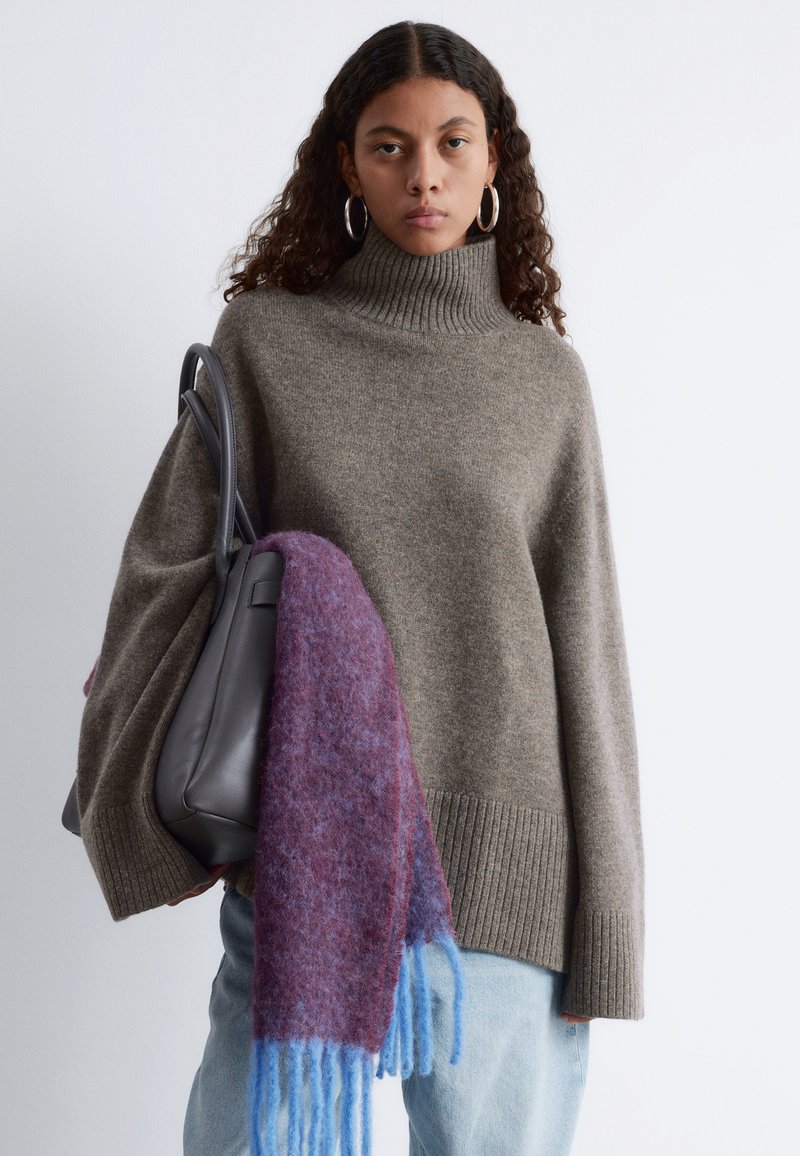 Beige übergroßer Rollkragenpullover mit gerippten Bündchen, kombiniert mit einem mehrfarbigen Schal in lila und blauen Tönen sowie einer schwarzen Handtasche.