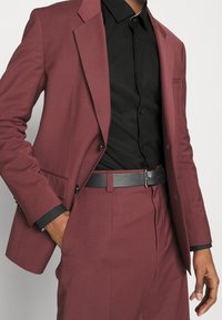 Giacca da abito maroon con camicia nera, caratterizzata da un design monopetto, rever a scollo, e un fazzoletto da taschino; abbinata a una cintura nera texturizzata.