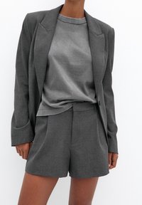 Blazer gris sur mesure avec col à revers pointu et t-shirt gris doux à col rond, associé à un short sur mesure assorti. Texture de tissu lisse, design épuré.