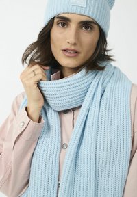 Bonnet en tricot bleu clair avec pompon et écharpe côtelée assortie, drapée doucement autour du cou, associée à une chemise rose clair.