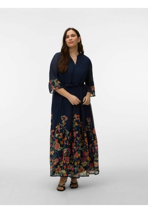 Mujer vistiendo un vestido maxi azul marino con estampado floral, mangas transparentes tres cuartos y cinturón en la cintura, posando frente a un fondo blanco sencillo.