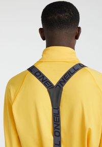 Suéter amarillo con alto cuello, que presenta correas de arnés grises con la marca "O'Neill" y un acento negro en la parte posterior. Textura suave.