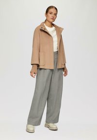 Beige Wollmantel mit hohem Kragen, Fronttaschen und Knopfakzenten. Kombiniert mit lockeren grauen Hosen und beigen Knöchelstiefeln. Glatte Textur.