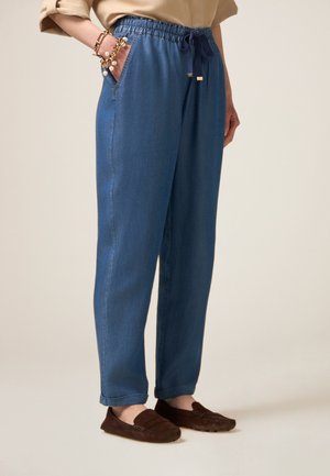 Comfortabele blauwe denimbroeken met een elastische tailleband, trekkoord, zijzakken en een ontspannen pasvorm. Gecombineerd met donkerbruine instap schoenen.
