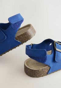 Blå mockaskor med korksål, utrustade med justerbara kardborreband och en texturerad yta för bra grepp. En enkel och praktisk design.