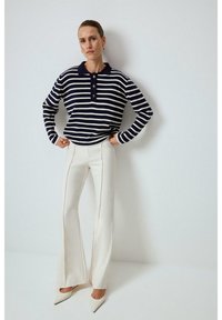 Randig marinblå och vit långärmad pikétröja med knappslå, kombinerad med krämfärgade utsvängda byxor och vita spetsiga klackskor.