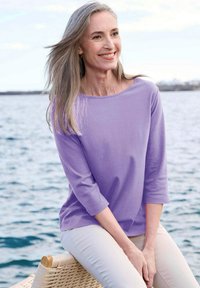 Lavendel 3/4 mouw top van zachte stof met een ontspannen pasvorm, gecombineerd met lichtbeige broek. Model zit bij water op een geweven stoel.