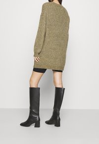 Olivgrüner, gestrickter Pullover mit gerippter Textur, langen Ärmeln und seitlichen Schlitzen, kombiniert mit kniehohen schwarzen Lederstiefeln mit Blockabsatz.