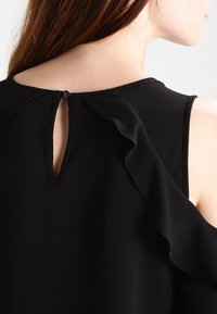 Svart blus med volangdetaljer vid axeln, nyckelhålsöppning bak och knäppning. Mjuk texturerad tyg, figurnära design.