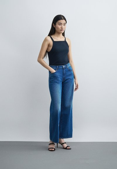 Γυναίκα με μακριά σκουρόχρωμα μαλλιά, που φοράει μαύρο αμάνικο τοπ, μπλε φαρδιά τζιν και μαύρα σανδάλια με τακούνι, στέκεται μπροστά σε έναν απλό τοίχο.