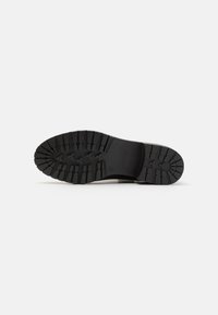 Semelle en caoutchouc noir avec des profondeurs prononcées et un motif strié. Présente un logo et un design robuste pour une traction et une durabilité accrues.