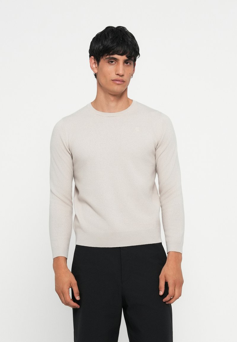 Suéter de malha em bege claro, com mangas longas, decote redondo e um discreto logótipo no peito, combinado com calças pretas.