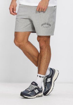 Mężczyzna w szarych szortach Calvin Klein, białych skarpetkach i ciemnoniebieskich butach New Balance, stojący ze skrzyżowanymi nogami na białej podłodze.