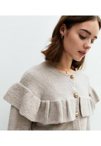 Beige stickad cardigan med volangdesign längs axlarna, gyllene knappar och en strukturerad yta.