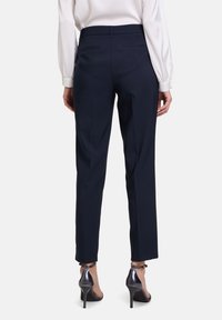 Pantaloni sartoriali blu navy con una vestibilità slim, dotati di tasche posteriori discrete e una texture liscia, abbinati a scarpe con tacco metallico.