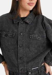 Donna che indossa una giacca in denim nero lavato con tasche a bottone e bottoni metallici, abbinata a pantaloni neri a vita alta coordinati.