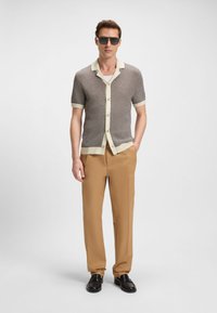 Cardigan a maniche corte in maglia marrone e crema con motivo a rilievo, bottoni e colletto a contrasto, abbinato a pantaloni beige e scarpe nere.