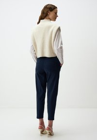 Un pull sans manches à côtes crème sur une chemise blanche à boutons, associé à un pantalon tapered bleu marine et des sandales à talons nude.