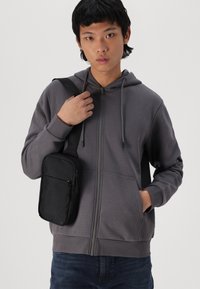 Felpa grigia con zip e cappuccio regolabile, dotata di tasche, e una borsa a tracolla nera con tasca frontale con zip, tenuta da una persona.