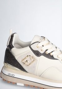 Sneaker bianco e marrone con dettagli in pelle e con motivi decorativi, dotato di accessori in oro e una suola testurizzata. Include lacci e una linguetta.