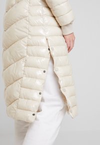 Abrigo tipo puffer beige con acabado brillante, que presenta un diseño acolchado y cierres de botones a presión a lo largo del dobladillo, mostrado con una parte superior blanca y pantalones.