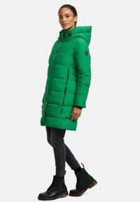 Abrigo acolchado verde con capucha, con cierre de cremallera y bolsillos laterales. Combinado con jeans ajustados negros y botas negras con cordones.