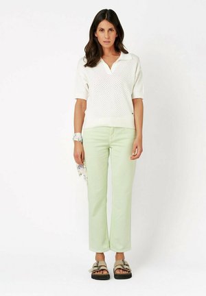 Woman standing wearing white knit short-sleeve top, light green pants, beige sandals, and holding a patterned scarf in her left hand.