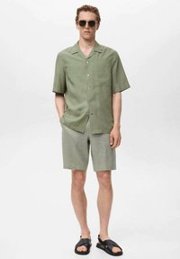 Cămașă cu mânecă scurtă, verde salvie, cu nasturi și un buzunar pe piept, asortată cu pantaloni scurți verzi deschis și sandale negre, purtată de un model masculin.