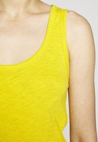 Keltainen toppi, jossa on hunajakennopääntie. Valmistettu pehmeästä kankaasta, jossa on ompeleet reunoilla, rento muotoilu ja sileä tekstuuri.