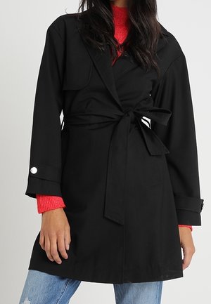 Frau trägt schwarzen Trenchcoat mit Gürtel über rotem Strickpullover und hellblauen, zerrissenen Jeans, stehend vor einfachem Hintergrund.