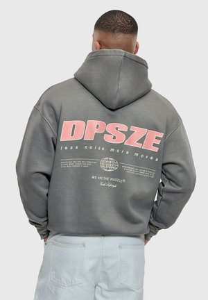 Man met kort haar die een grijze hoodie draagt met roze tekst "DPSZE less noise more moves" en lichte spijkerbroek, met de rug naar de camera toe.