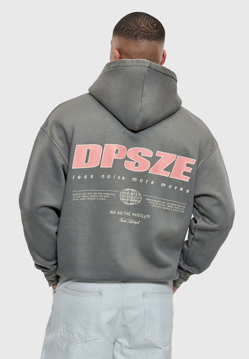 Homme aux cheveux courts portant un sweat à capuche gris avec le texte rose "DPSZE less noise more moves" et un pantalon en jean clair, tourné de dos à la caméra.