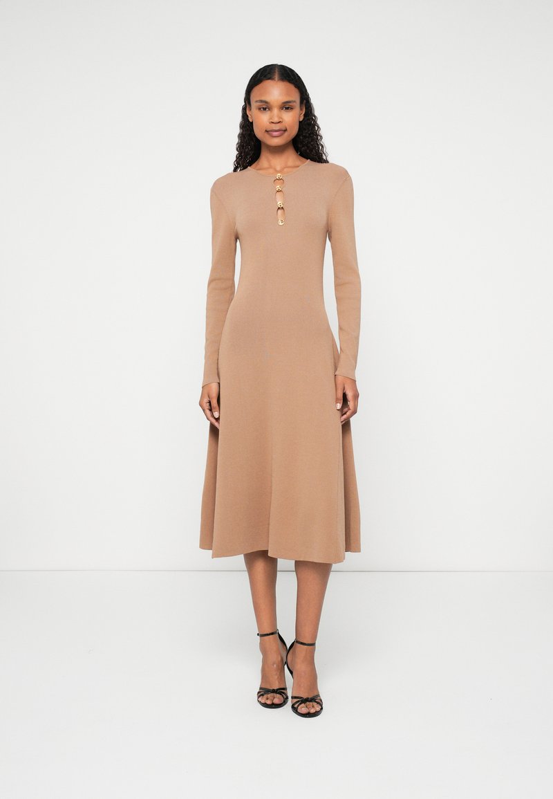 Világosbarna kötött ruha hosszú ujjal, által térd középig érő hosszúságban, gombos díszítéssel a nyakvonalon, fekete, pántos magassarkúval viselve.