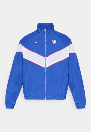 Casaco de nylon azul com riscas diagonais brancas e vermelhas, fecho com zíper, gola alta e logótipo bordado no peito. Punhos e cintura elásticos.