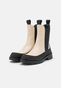 Bottines beige et noires avec un dessus brillant, une semelle en caoutchouc épaisse, des panneaux latéraux élastiques et une finition texturée.