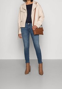 Beige läderbikerjacka med dragkedjor, marinblå topp, blå skinnyjeans och bruna klackade ankelstövlar. Brun axelväska med vävd detalj.