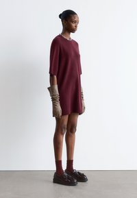 Burgunderfarbenes gestricktes oversized Kleid mit kurzen Ärmeln, kombiniert mit braunen gerafften Handschuhen und dunklen Slippern, ergänzt durch burgunderfarbene Socken.