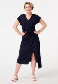 Marineblauwe wikkeljurk met een V-hals, korte mouwen, gedraaide details bij de taille en een zijsplit. Gecombineerd met lichtgekleurde blokhak sandalen.