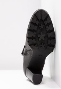 Botín negro de cuero con suela de goma texturizada, que presenta un tacón cuadrado y cremalleras decorativas a lo largo de los laterales.