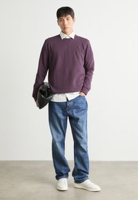 Lilla genser med rund hals over en hvit skjorte med krage, kombinert med blå jeans og hvite sneakers. Holder en svart veske. Enkelt bakgrunn.
