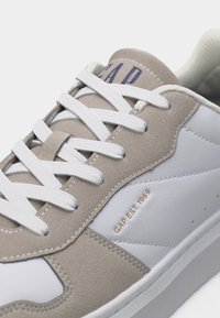 Άσπρο και μπεζ καθημερινό αθλητικό παπούτσι με άσπρα κορδόνια και χρυσό κείμενο "GAP EST. 1969" στην πλαϊνή επιφάνεια.