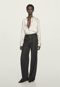 Satin, cremefarvet skjorte med dyb V-udskæring; parret med højtaljede, mørkegrå vide bukser. Enkel sølv halskæde som accent.