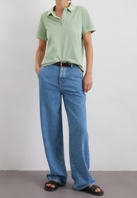 Henkilö, joka on pukeutunut vaaleanvihreään lyhythihaiseen polo-paitaan, löysiin sinisiin farkkuihin, mustaan vyöhön ja mustiin avointoiseen sandaaleihin, seisoo valkoista seinää vasten.