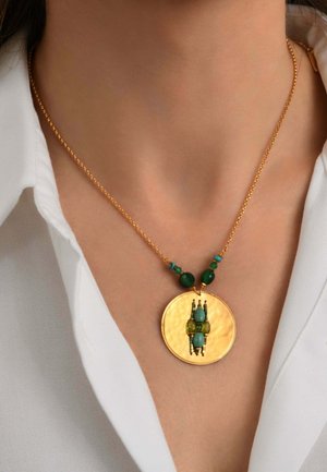Collier chaîne en or avec pendentif disque en or martelé décoré de perles vertes et turquoise porté par une personne avec une chemise blanche.