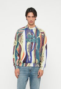 Farverig strikket sweater med abstrakte mønstre i grøn, lilla og orange. Har en rund hals og ribbede manchetter og kant. Frontlogo i blå.