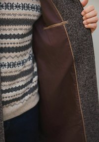 Inimene, kes kannab mustriga sviitrit ja tumedaid teksaseid, hoiab lahti pruuni tweedist jope, näidates selle siledat pruuni sisevoodrit ja taskut.